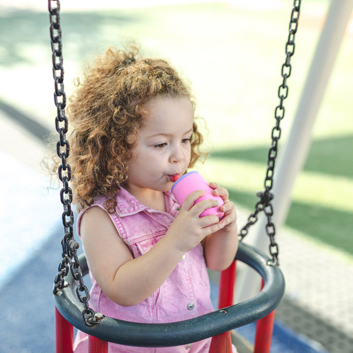 A young child with curly hair sits on a swing at a playground, wearing a pink sleeveless shirt and sipping from a straw in a pink cup. The background features green grass and colorful playground equipment.