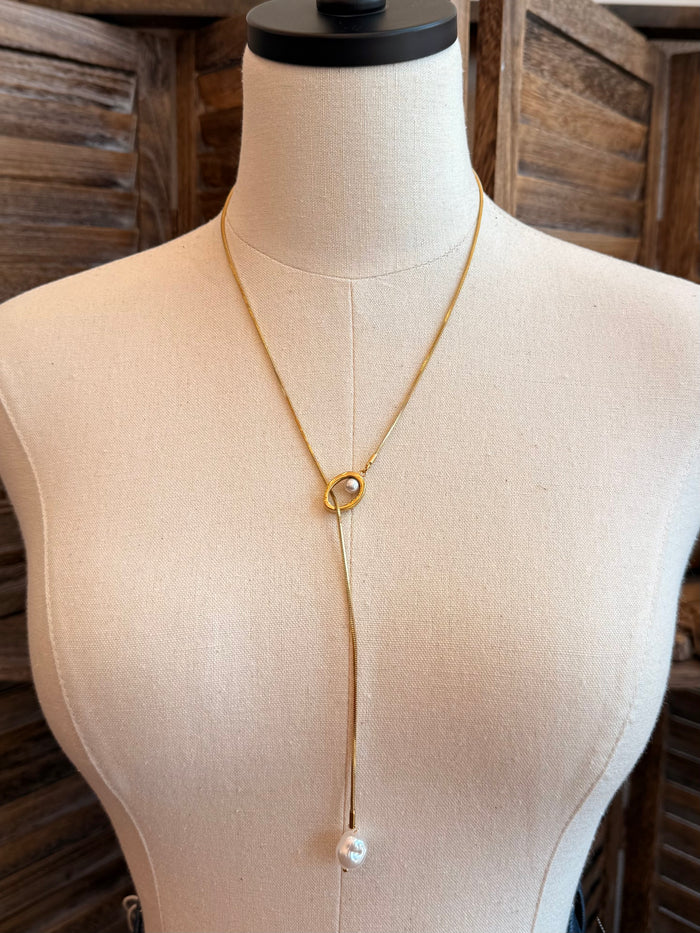 Gold lariat necklace with round clasp and single pearl drop, draped on mannequin