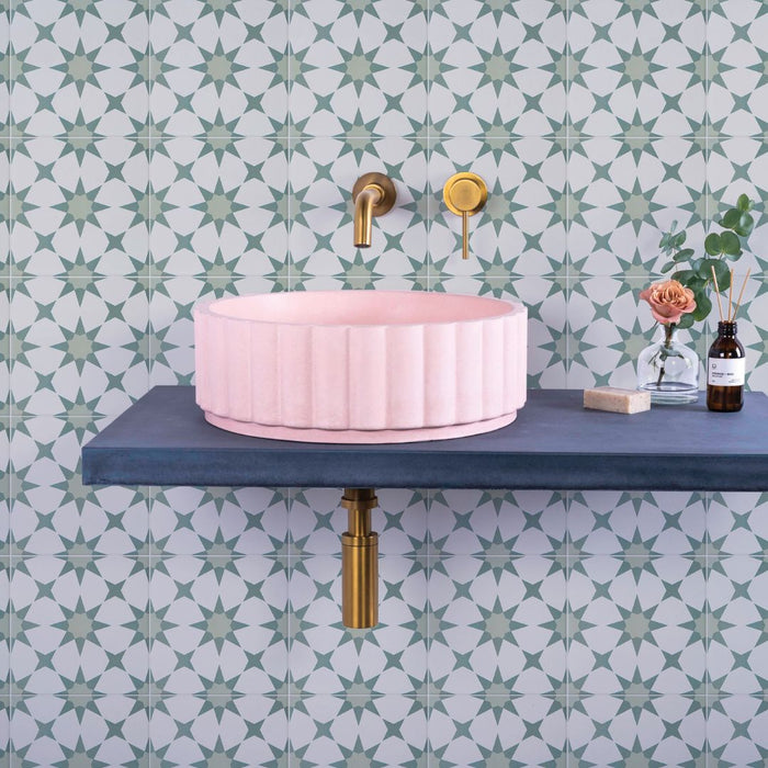 Pink sink on a blue shelf with gold fixtures against green patterned wall tiles