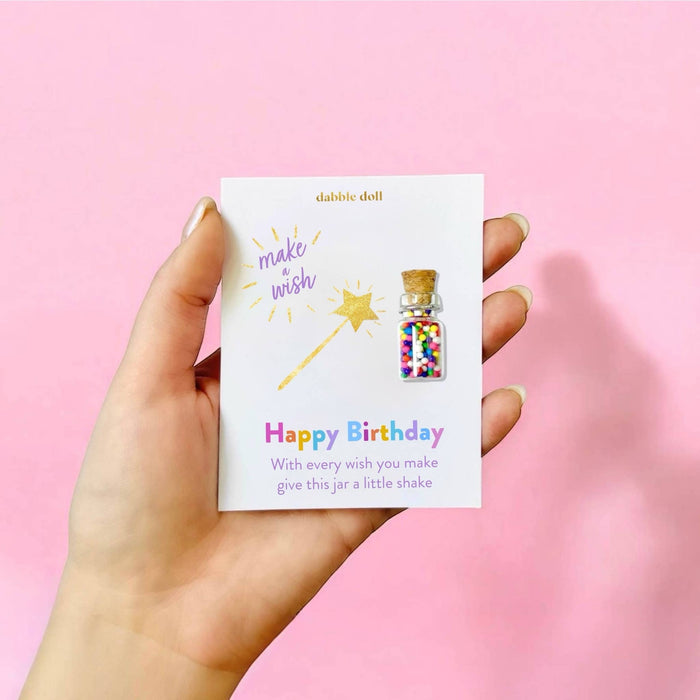 A hand holds a white card featuring a small birthday gift: a glittery bottle pin with colorful confetti and a gold star wand illustration. The card reads 'dabble doll' and 'Happy Birthday'.