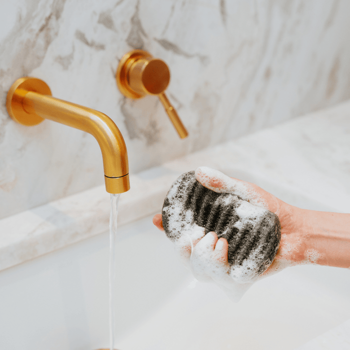 Hand holding a soapy Konjac Bamboo Charcoal Body Sponge | Bathorium under running water from a modern gold faucet in a marble bathroom sink for skincare and body care wellness.