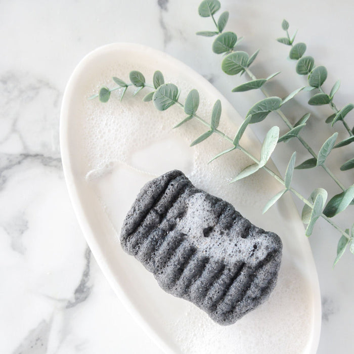 Konjac Bamboo Charcoal Body Sponge | Bathorium resting on a white soap dish with foamy bubbles and eucalyptus branches on a marble surface, highlighting natural skincare and body care.