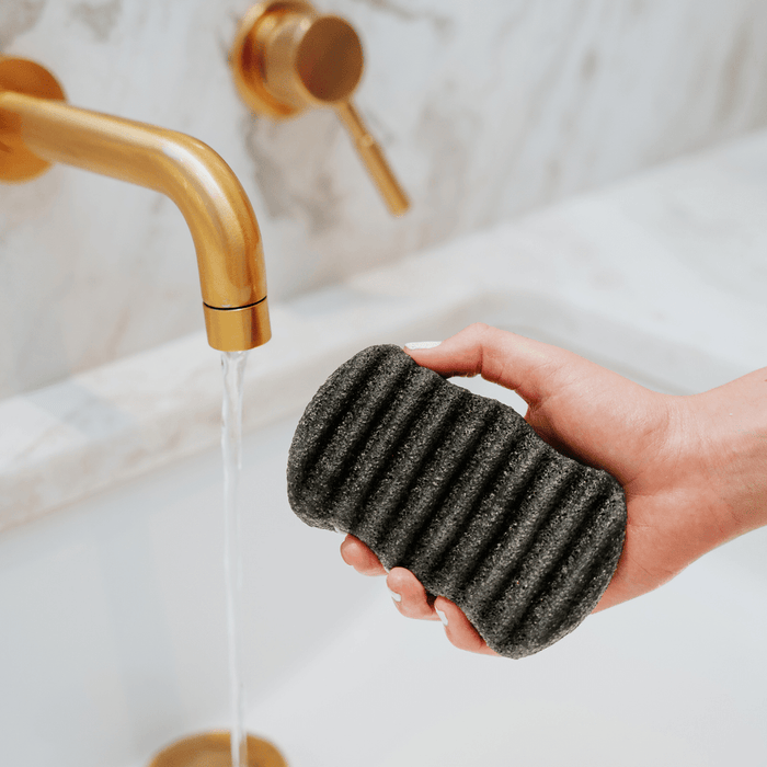 Hand holding a black textured Konjac Bamboo Charcoal Body Sponge | Bathorium under running water from a modern gold faucet over a white marble sink for skincare and body care routine.
