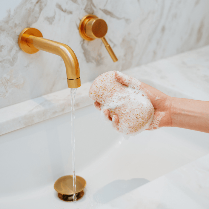 Hand holding a foamy Konjac Walnut Exfoliating Body Sponge | Bathorium under running water from a gold faucet in a marble bathroom sink, used for skincare and body care.