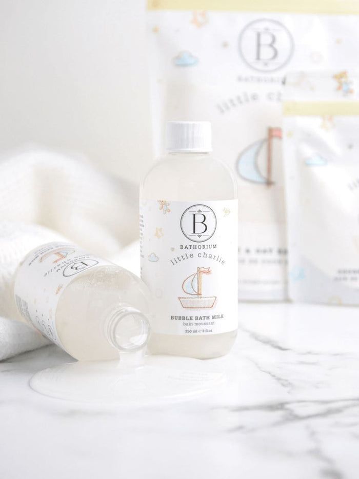 Two bottles of little charlie™ Bubble Bath Milk | Bathorium on a white marble surface, one standing upright and one lying down with liquid spilled beside it, surrounded by soft towels and blurred packaging in the background.