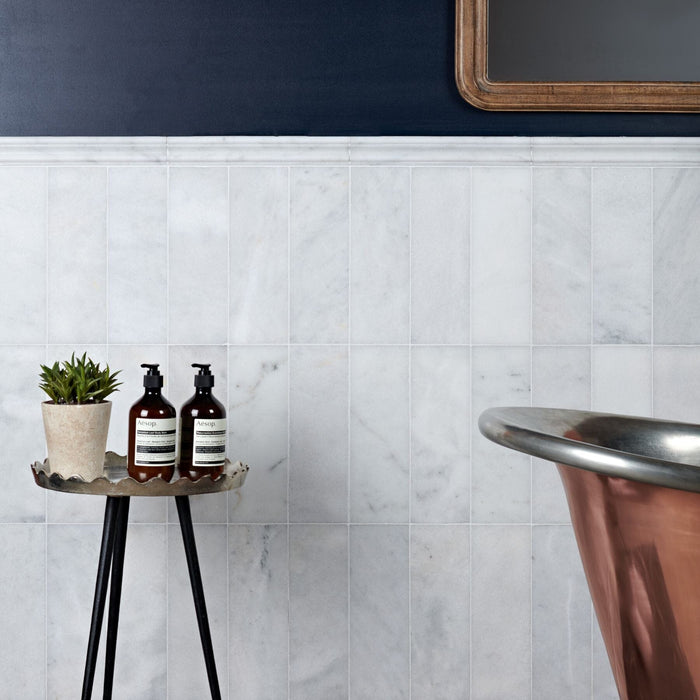 Bathroom setting with a small table holding bottles and a plant, against a marble tiled wall.