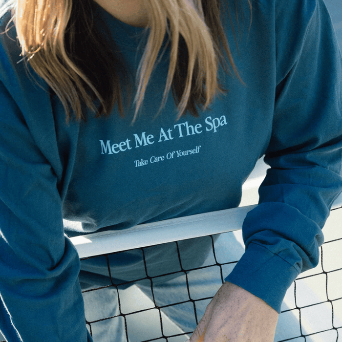 Person wearing a blue long sleeve shirt with the text "Meet Me At The Spa - Take Care Of Yourself Long Sleeve T-Shirt | Lucky Owl" leaning on a tennis net, promoting wellness and self-care.