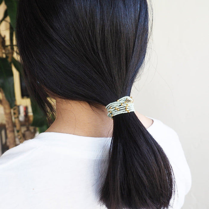 Woman with dark hair styled in a low ponytail secured by the Wonder Wrap Bracelet (White Pearl & Gold Multi) | Love You More, featuring multiple strands of white pearls and gold accents.